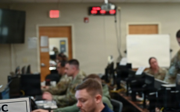 The image shows personnel working inside an Emergency Operations Center (EOC), as indicated by a nameplate reading 'EOC Director' in the foreground. Multiple individuals in military uniforms (camouflage) and civilian attire are seated at workstations with computers. The setting appears to be an active operations or exercise environment with monitoring equipment and displays visible.