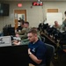 The image shows personnel working inside an Emergency Operations Center (EOC), as indicated by a nameplate reading 'EOC Director' in the foreground. Multiple individuals in military uniforms (camouflage) and civilian attire are seated at workstations with computers. The setting appears to be an active operations or exercise environment with monitoring equipment and displays visible.