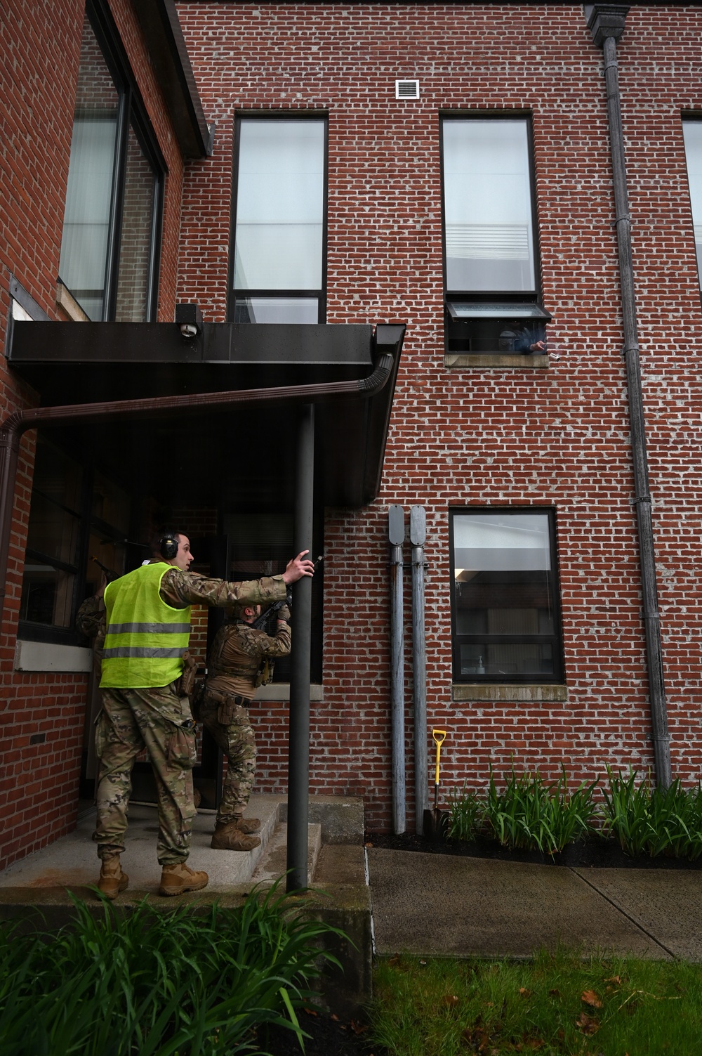 Two personnel in military camouflage uniforms are conducting what appears to be a tactical building entry exercise at a red brick building. One individual wears a bright yellow/green safety vest and hearing protection, suggesting a training or observer role, while the other appears to be making entry through a doorway. A third person is partially visible leaning out of a second-floor window.