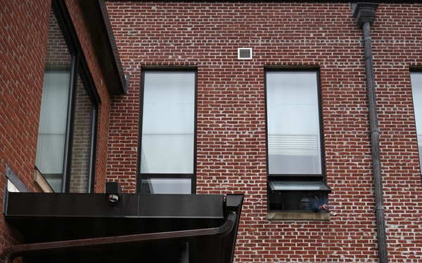 Two personnel in military camouflage uniforms are conducting what appears to be a tactical building entry exercise at a red brick building. One individual wears a bright yellow/green safety vest and hearing protection, suggesting a training or observer role, while the other appears to be making entry through a doorway. A third person is partially visible leaning out of a second-floor window.