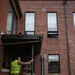 Two personnel in military camouflage uniforms are conducting what appears to be a tactical building entry exercise at a red brick building. One individual wears a bright yellow/green safety vest and hearing protection, suggesting a training or observer role, while the other appears to be making entry through a doorway. A third person is partially visible leaning out of a second-floor window.
