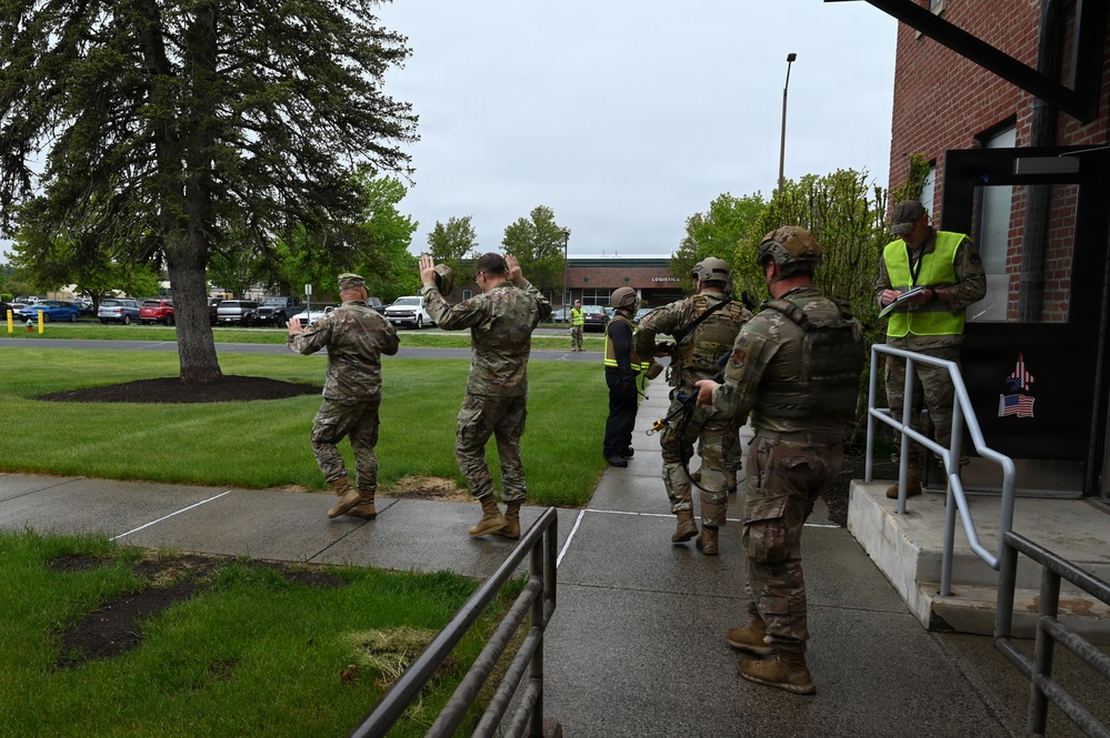 The image shows a group of military personnel in camouflage uniforms participating in what appears to be a training exercise or drill outside a brick building. Two individuals in the foreground have their hands raised, while others in tactical gear line up near building entrance steps. A civilian observer in a high-visibility yellow vest appears to be monitoring and documenting the exercise.