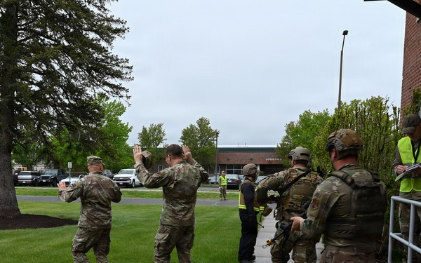 The image shows a group of military personnel in camouflage uniforms participating in what appears to be a training exercise or drill outside a brick building. Two individuals in the foreground have their hands raised, while others in tactical gear line up near building entrance steps. A civilian observer in a high-visibility yellow vest appears to be monitoring and documenting the exercise.