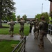 The image shows a group of military personnel in camouflage uniforms participating in what appears to be a training exercise or drill outside a brick building. Two individuals in the foreground have their hands raised, while others in tactical gear line up near building entrance steps. A civilian observer in a high-visibility yellow vest appears to be monitoring and documenting the exercise.