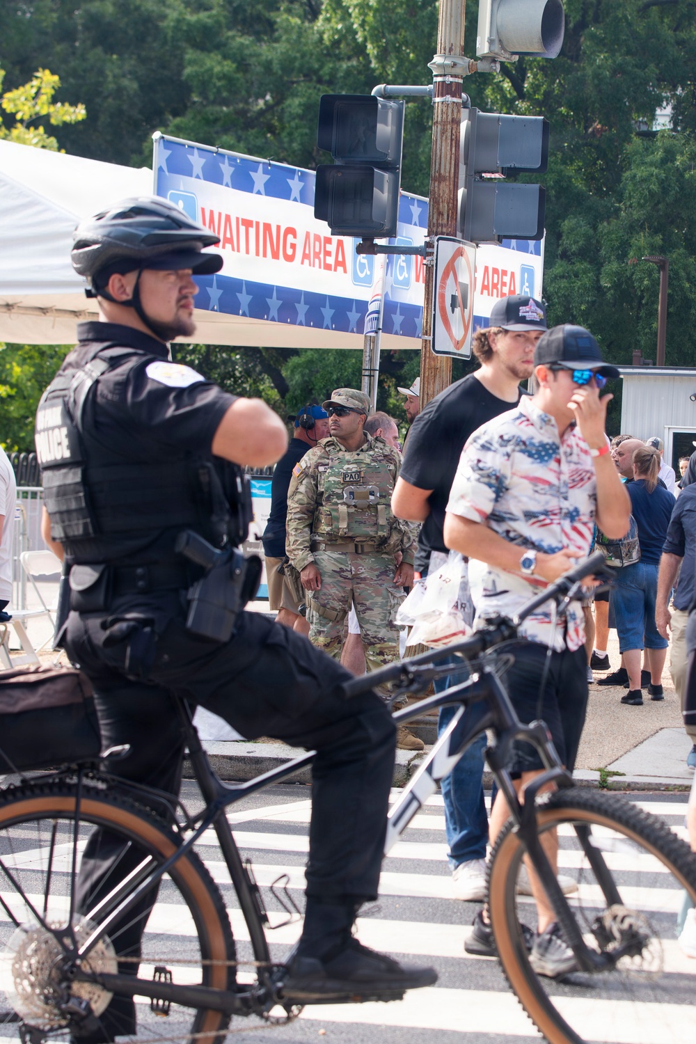 A public outdoor event with visible security presence, including a bicycle police officer in full tactical gear and a U.S. Army soldier in camouflage uniform with a 'PAO' (Public Affairs Officer) patch. A 'Waiting Area' banner with accessibility symbols is displayed on a traffic signal pole, suggesting an organized public gathering. Civilians in casual attire, including patriotic-themed clothing, are present in the background.