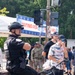 A public outdoor event with visible security presence, including a bicycle police officer in full tactical gear and a U.S. Army soldier in camouflage uniform with a 'PAO' (Public Affairs Officer) patch. A 'Waiting Area' banner with accessibility symbols is displayed on a traffic signal pole, suggesting an organized public gathering. Civilians in casual attire, including patriotic-themed clothing, are present in the background.