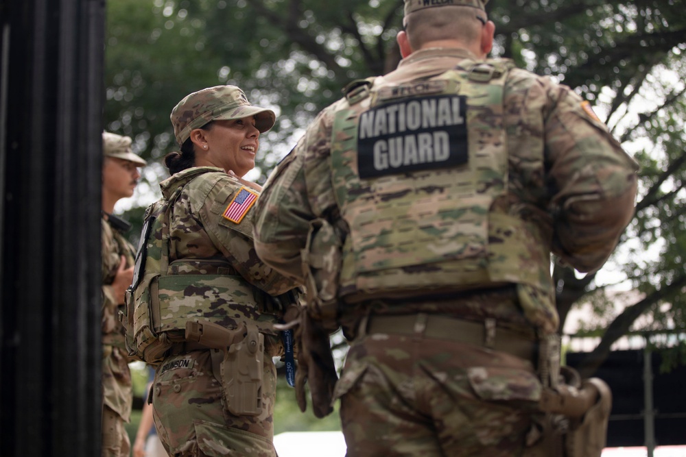 The image shows U.S. National Guard soldiers in military camouflage (OCP/MultiCam) uniforms engaged in conversation outdoors. A female soldier with the name tape 'BRUNSON' and a U.S. flag patch is smiling while speaking with a male soldier whose back is turned, displaying a 'NATIONAL GUARD' patch. The setting appears to be an outdoor area with trees in the background.