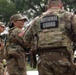 The image shows U.S. National Guard soldiers in military camouflage (OCP/MultiCam) uniforms engaged in conversation outdoors. A female soldier with the name tape 'BRUNSON' and a U.S. flag patch is smiling while speaking with a male soldier whose back is turned, displaying a 'NATIONAL GUARD' patch. The setting appears to be an outdoor area with trees in the background.