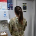 A person wearing US Army OCP (Operational Camouflage Pattern) uniform is standing with their back to the camera in what appears to be a medical or administrative facility. They are facing a wall with posted notices and a first aid station, standing near a counter with paperwork and supplies. The setting suggests a military medical or health clinic environment.