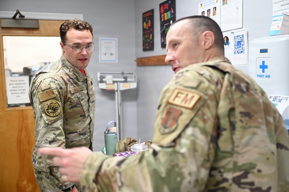 Two military personnel in OCP (Operational Camouflage Pattern) uniforms are engaged in conversation inside what appears to be a medical or fitness facility. The individual facing the camera wears glasses and has a unit patch with a US flag on their left shoulder. The other individual, seen from behind, displays an 'FM' (Flight Medicine or Force Management) patch and an Air Force symbol patch.
