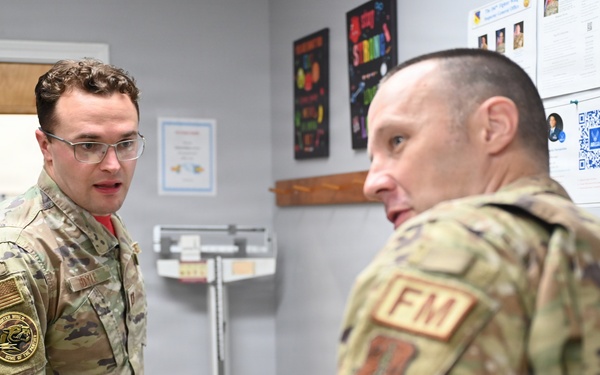 Two military personnel in OCP (Operational Camouflage Pattern) uniforms are engaged in conversation inside what appears to be a medical or fitness facility. The individual facing the camera wears glasses and has a unit patch with a US flag on their left shoulder. The other individual, seen from behind, displays an 'FM' (Flight Medicine or Force Management) patch and an Air Force symbol patch.