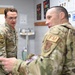 Two military personnel in OCP (Operational Camouflage Pattern) uniforms are engaged in conversation inside what appears to be a medical or fitness facility. The individual facing the camera wears glasses and has a unit patch with a US flag on their left shoulder. The other individual, seen from behind, displays an 'FM' (Flight Medicine or Force Management) patch and an Air Force symbol patch.