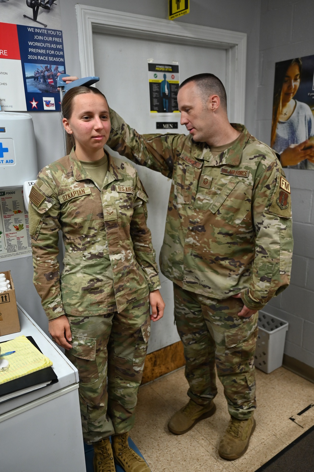 Two U.S. Air Force personnel in OCP (Operational Camouflage Pattern) uniforms are conducting a height measurement at what appears to be a fitness center. The male airman is using a measuring device on top of the female airman's head while she stands against a wall. The setting appears to be a military fitness facility, likely during a physical fitness assessment.