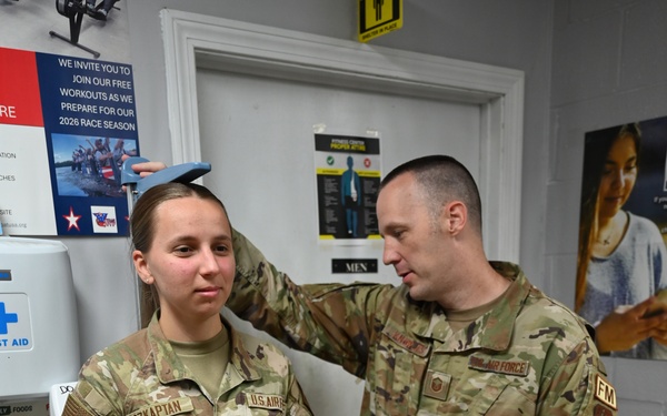 Two U.S. Air Force personnel in OCP (Operational Camouflage Pattern) uniforms are conducting a height measurement at what appears to be a fitness center. The male airman is using a measuring device on top of the female airman's head while she stands against a wall. The setting appears to be a military fitness facility, likely during a physical fitness assessment.