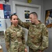 Two U.S. Air Force personnel in OCP (Operational Camouflage Pattern) uniforms are conducting a height measurement at what appears to be a fitness center. The male airman is using a measuring device on top of the female airman's head while she stands against a wall. The setting appears to be a military fitness facility, likely during a physical fitness assessment.