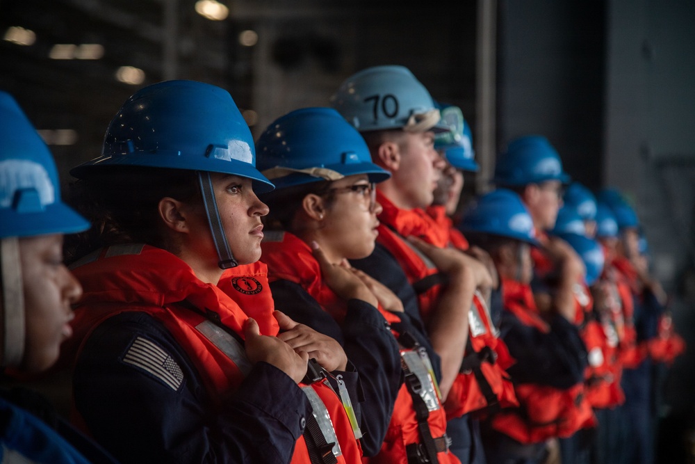 A group of personnel wearing blue hard hats and orange life vests over navy blue uniforms are standing in a line, appearing to be at attention or receiving instruction. One helmet is visibly marked with the number '70'. The setting appears to be an industrial or naval vessel interior, suggesting a safety drill, muster, or orientation exercise.
