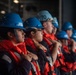 A group of personnel wearing blue hard hats and orange life vests over navy blue uniforms are standing in a line, appearing to be at attention or receiving instruction. One helmet is visibly marked with the number '70'. The setting appears to be an industrial or naval vessel interior, suggesting a safety drill, muster, or orientation exercise.