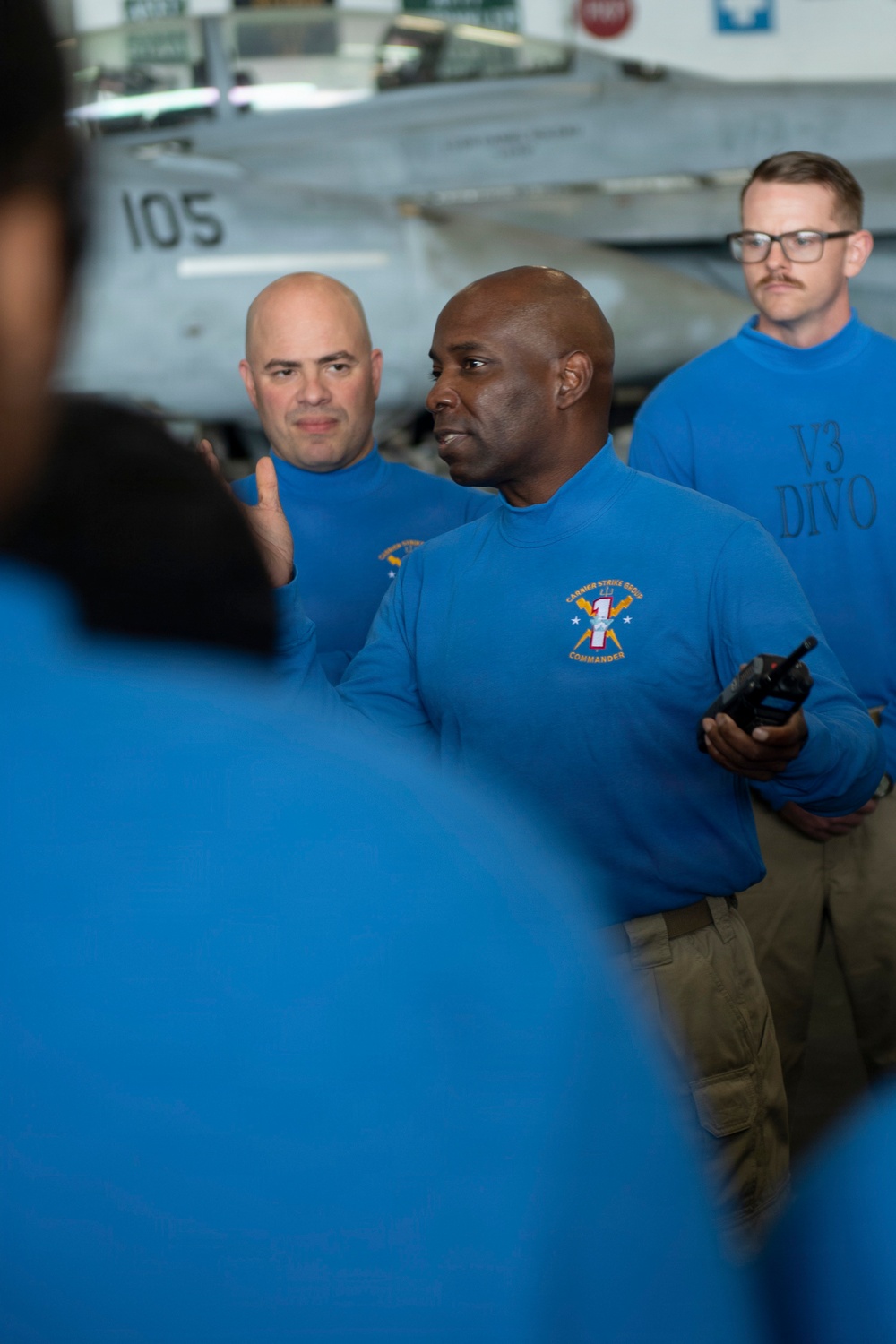 Several individuals wearing blue long-sleeve shirts are gathered in what appears to be an aircraft carrier hangar bay, with a military jet aircraft (numbered 105) visible in the background. The central figure, wearing a shirt with a 'Carrier Strike Group Commander' patch, appears to be addressing a group while holding a radio. A third person in the background wears a shirt labeled 'V3 DIVO', indicating a divisional officer role.