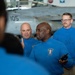Several individuals wearing blue long-sleeve shirts are gathered in what appears to be an aircraft carrier hangar bay, with a military jet aircraft (numbered 105) visible in the background. The central figure, wearing a shirt with a 'Carrier Strike Group Commander' patch, appears to be addressing a group while holding a radio. A third person in the background wears a shirt labeled 'V3 DIVO', indicating a divisional officer role.