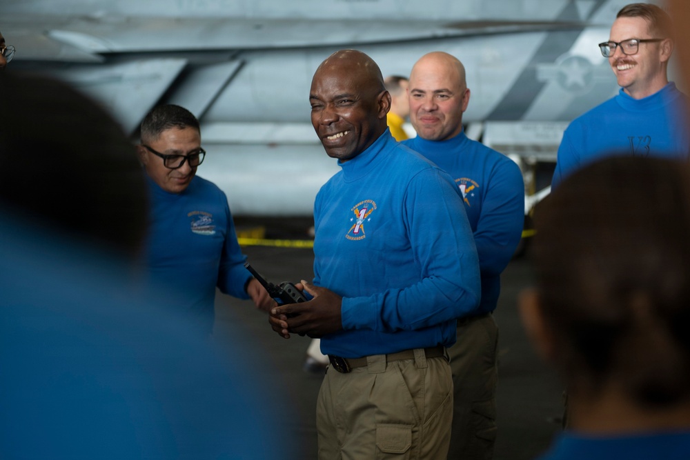A group of individuals wearing matching blue uniform shirts are gathered in what appears to be an aircraft hangar bay, likely aboard a naval vessel. The central figure, a tall man holding a radio/walkie-talkie, is smiling broadly while addressing the group. A military aircraft is visible in the background.
