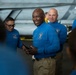 A group of individuals wearing matching blue uniform shirts are gathered in what appears to be an aircraft hangar bay, likely aboard a naval vessel. The central figure, a tall man holding a radio/walkie-talkie, is smiling broadly while addressing the group. A military aircraft is visible in the background.