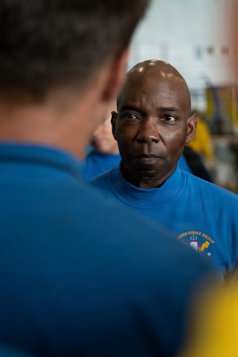 The image shows two individuals in blue shirts, with one man in sharp focus facing the camera and another person seen from behind in the foreground. The man in focus is bald, wearing a blue shirt with a partially visible 'Carrier Strike Group' logo. The setting appears to be an indoor facility, possibly a military or naval environment.
