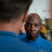 The image shows two individuals in blue shirts, with one man in sharp focus facing the camera and another person seen from behind in the foreground. The man in focus is bald, wearing a blue shirt with a partially visible 'Carrier Strike Group' logo. The setting appears to be an indoor facility, possibly a military or naval environment.