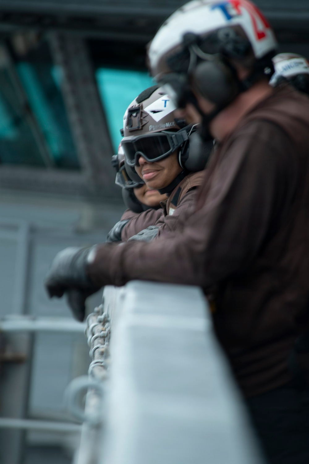 Two military personnel, likely U.S. Navy sailors, are leaning against a railing on what appears to be the deck of a naval vessel. Both are wearing brown flight deck jerseys, helmets with hearing protection, protective goggles, and black gloves. The person in the background is smiling and has a helmet marked with the letter 'T' and a name tag.