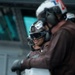 Two military personnel, likely U.S. Navy sailors, are leaning against a railing on what appears to be the deck of a naval vessel. Both are wearing brown flight deck jerseys, helmets with hearing protection, protective goggles, and black gloves. The person in the background is smiling and has a helmet marked with the letter 'T' and a name tag.
