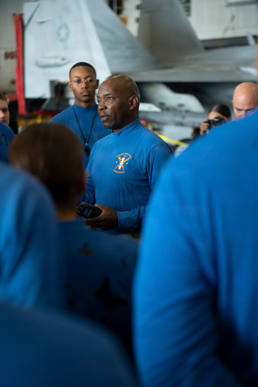 A group of personnel wearing matching blue long-sleeve shirts are gathered in what appears to be an aircraft carrier hangar bay. The central figure, whose shirt bears an emblem with the word 'COMMANDER,' appears to be addressing the group while holding a handheld device. Military aircraft are visible in the background.