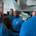 A group of personnel wearing matching blue long-sleeve shirts are gathered in what appears to be an aircraft carrier hangar bay. The central figure, whose shirt bears an emblem with the word 'COMMANDER,' appears to be addressing the group while holding a handheld device. Military aircraft are visible in the background.