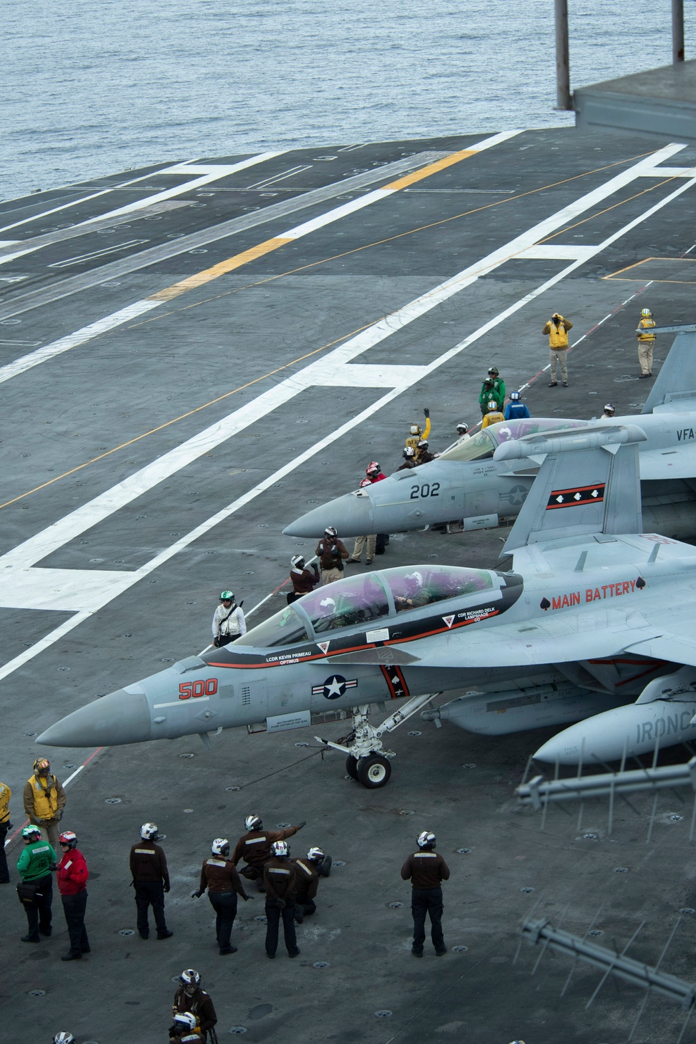 The image shows a U.S. Navy aircraft carrier flight deck with multiple F/A-18 Super Hornet jets staged for operations. The foreground aircraft is numbered '500' and marked 'MAIN BATTERY,' while a second aircraft numbered '202' is visible behind it. Numerous flight deck crew members in color-coded jerseys are visible performing pre-launch or marshaling duties.