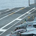 The image shows a U.S. Navy aircraft carrier flight deck with multiple F/A-18 Super Hornet jets staged for operations. The foreground aircraft is numbered '500' and marked 'MAIN BATTERY,' while a second aircraft numbered '202' is visible behind it. Numerous flight deck crew members in color-coded jerseys are visible performing pre-launch or marshaling duties.