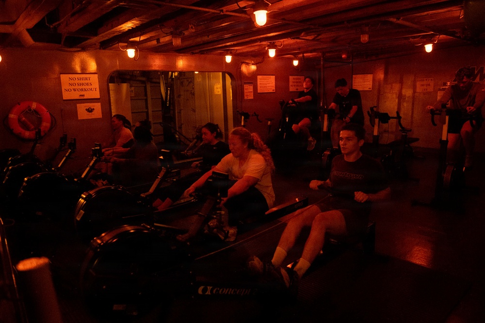 A group of people are working out in a dimly lit gym space with warm red/orange ambient lighting. Multiple individuals are using Concept2 rowing machines in what appears to be a confined, low-ceiling facility, possibly aboard a ship or in a basement gym. A sign on the wall reads 'NO SHIRT, NO SHOES, NO WORKOUT' and a life preserver ring is visible, suggesting a nautical or maritime setting.