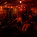 A group of people are working out in a dimly lit gym space with warm red/orange ambient lighting. Multiple individuals are using Concept2 rowing machines in what appears to be a confined, low-ceiling facility, possibly aboard a ship or in a basement gym. A sign on the wall reads 'NO SHIRT, NO SHOES, NO WORKOUT' and a life preserver ring is visible, suggesting a nautical or maritime setting.