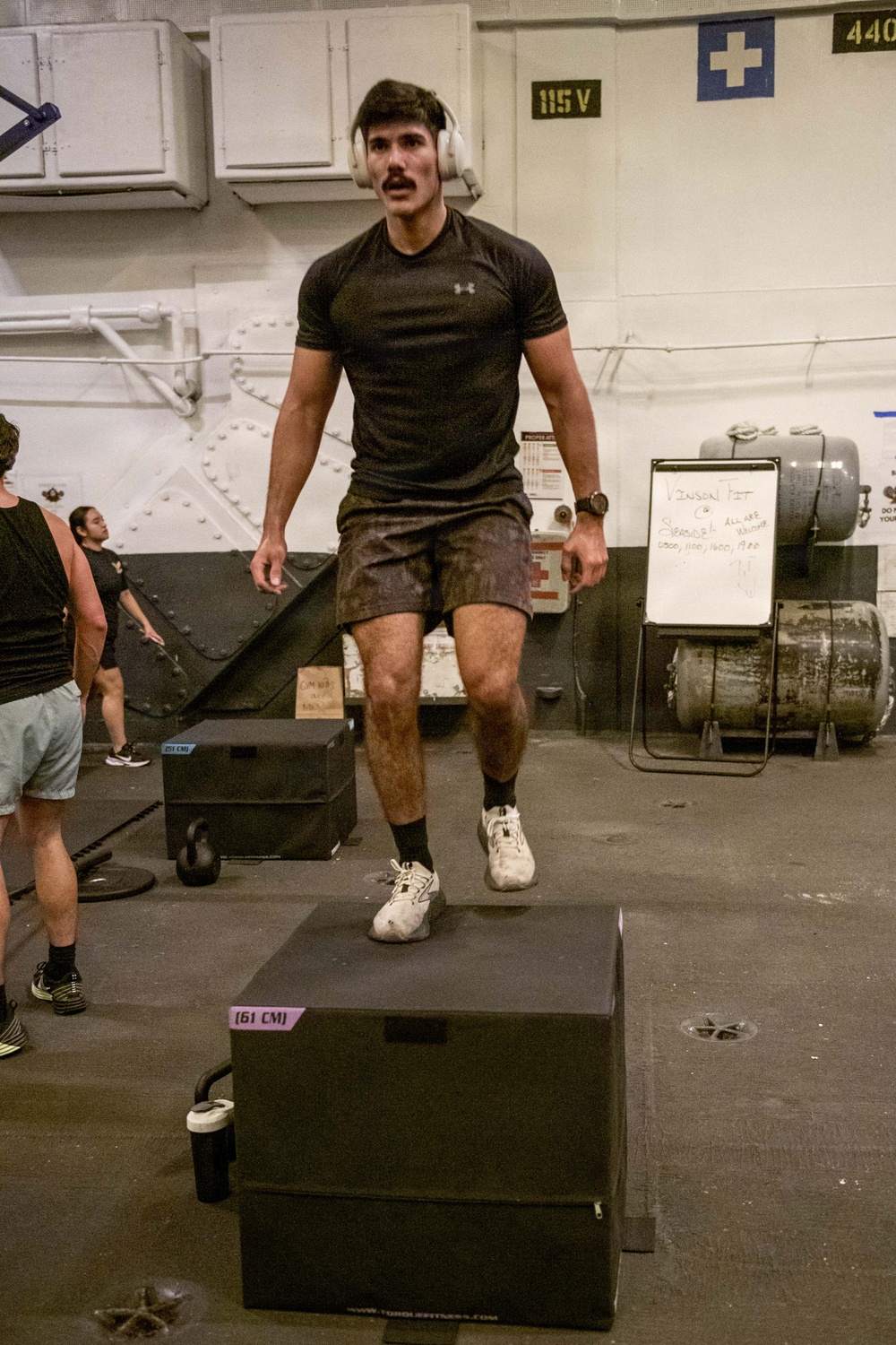 A young man wearing over-ear headphones and an Under Armour t-shirt is performing a box jump exercise onto a 61 cm plyo box in what appears to be a military or industrial-style gym. The facility contains various fitness equipment including kettlebells, additional plyo boxes, and a whiteboard with workout information. The setting appears to be aboard a naval vessel or military installation based on the riveted steel walls and electrical signage.