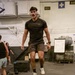 A young man wearing over-ear headphones and an Under Armour t-shirt is performing a box jump exercise onto a 61 cm plyo box in what appears to be a military or industrial-style gym. The facility contains various fitness equipment including kettlebells, additional plyo boxes, and a whiteboard with workout information. The setting appears to be aboard a naval vessel or military installation based on the riveted steel walls and electrical signage.