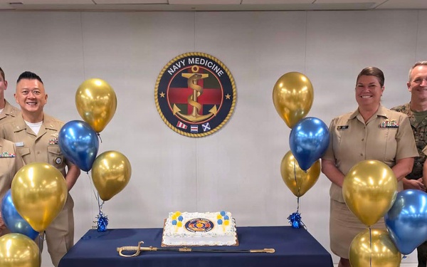 A group of U.S. Navy Medicine personnel pose for a celebratory photo in front of a Navy Medicine emblem/seal. The occasion appears to be an anniversary or birthday celebration, evidenced by a decorated cake on a navy blue draped table and gold and blue balloons. Personnel are wearing a mix of Navy Service Uniform (khaki) and Navy Working Uniform (NWU digital camouflage).
