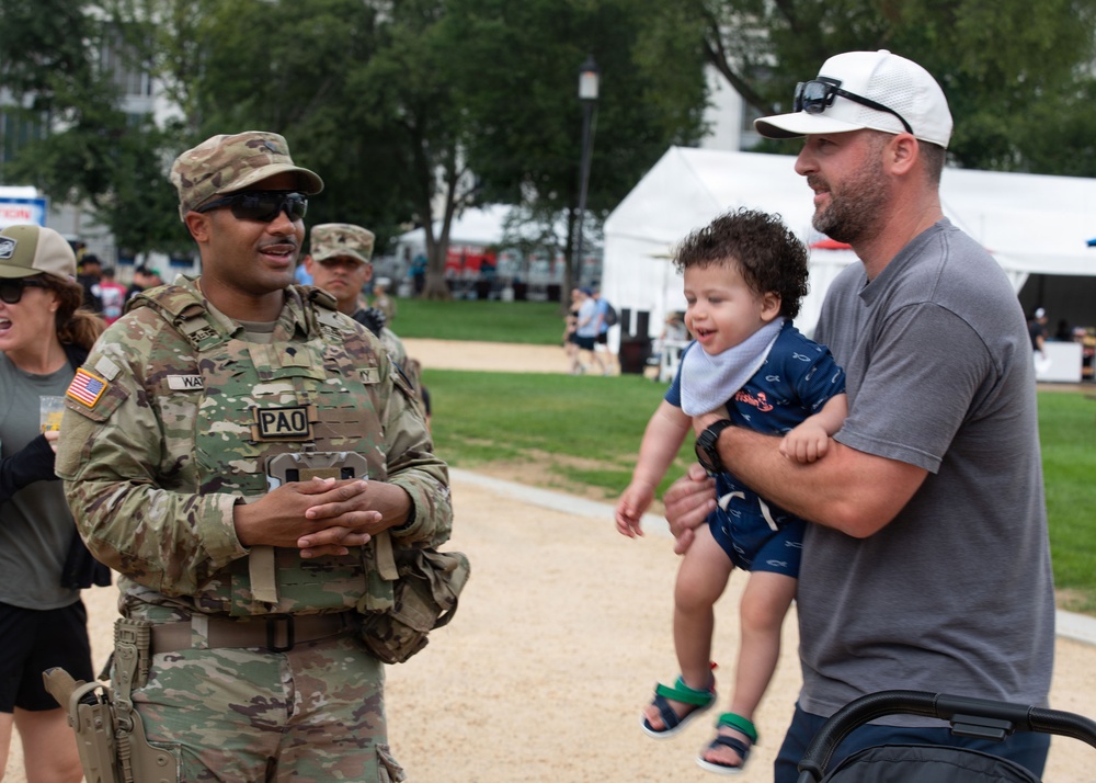 A U.S. Army soldier in full OCP (Operational Camouflage Pattern) uniform with a 'PAO' (Public Affairs Officer) chest marker is engaged in a friendly conversation with a civilian man holding a young toddler at what appears to be an outdoor public event. The interaction appears warm and casual, with the baby appearing animated and cheerful. The setting suggests a community or military public outreach event held in a park-like area.