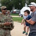A U.S. Army soldier in full OCP (Operational Camouflage Pattern) uniform with a 'PAO' (Public Affairs Officer) chest marker is engaged in a friendly conversation with a civilian man holding a young toddler at what appears to be an outdoor public event. The interaction appears warm and casual, with the baby appearing animated and cheerful. The setting suggests a community or military public outreach event held in a park-like area.
