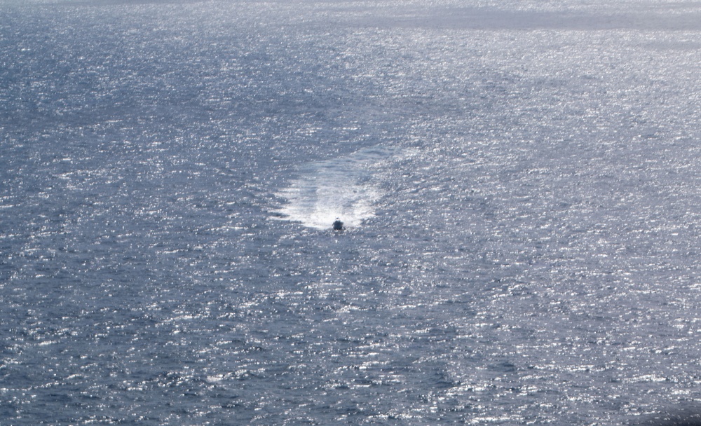 An aerial or elevated view of an open ocean with a single small vessel visible near the center of the frame. The water surface is heavily textured with sunlight glinting off the waves, creating a silver-blue shimmering effect. The boat appears to be in motion, leaving a visible wake trail behind it.