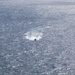 An aerial or elevated view of an open ocean with a single small vessel visible near the center of the frame. The water surface is heavily textured with sunlight glinting off the waves, creating a silver-blue shimmering effect. The boat appears to be in motion, leaving a visible wake trail behind it.