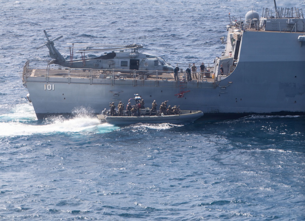 A military naval operation is underway showing a large gray warship marked '101' with a helicopter numbered '714' on its deck. A rigid inflatable boat (RIB) carrying multiple armed personnel in tactical gear is operating alongside the vessel. This appears to be a maritime special operations or boarding team exercise/deployment.