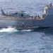 A military naval operation is underway showing a large gray warship marked '101' with a helicopter numbered '714' on its deck. A rigid inflatable boat (RIB) carrying multiple armed personnel in tactical gear is operating alongside the vessel. This appears to be a maritime special operations or boarding team exercise/deployment.
