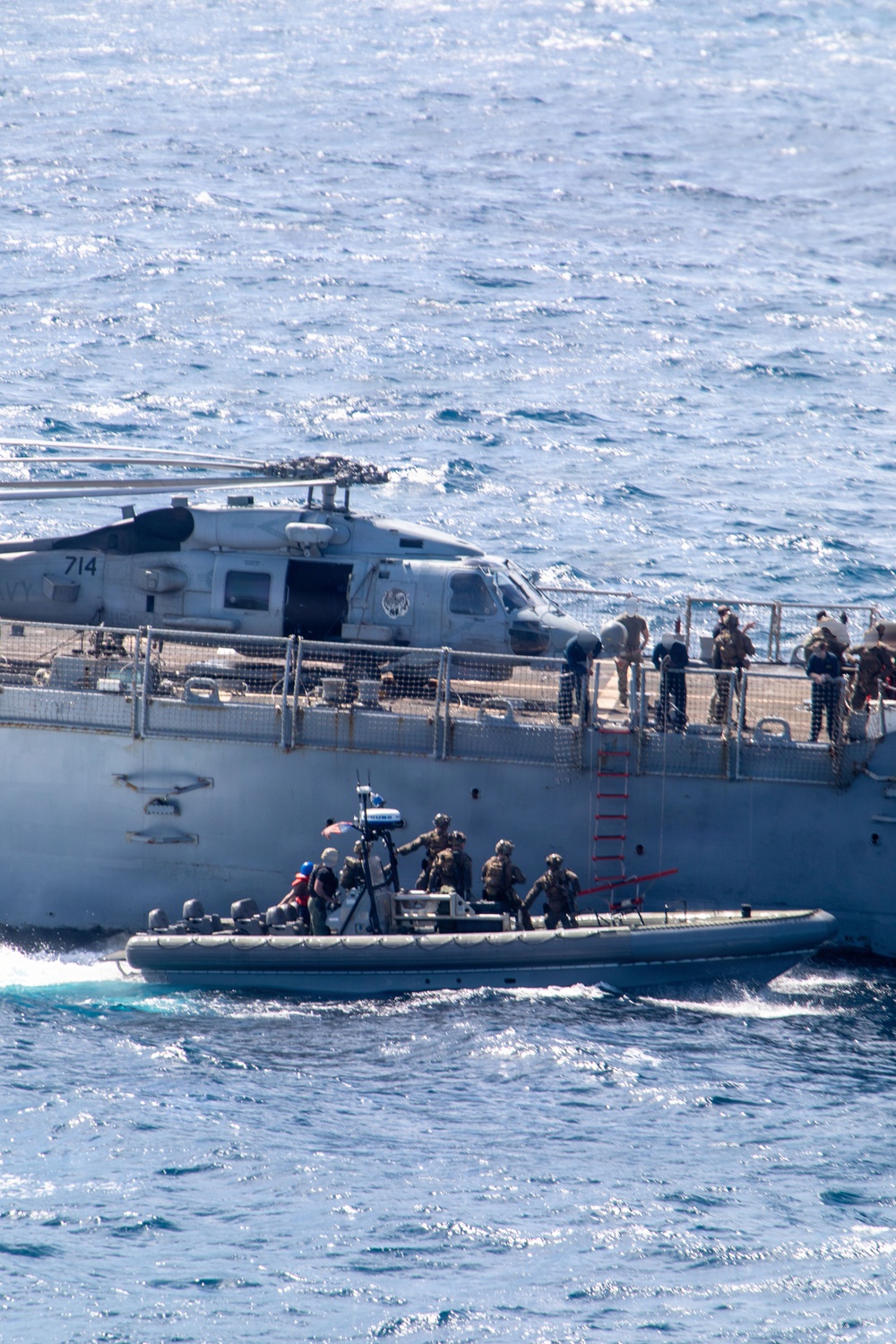 A military maritime operation is underway showing a rigid-hull inflatable boat (RHIB) with armed personnel alongside a larger naval vessel. A military helicopter marked '714' is positioned on the deck of the larger ship, with crew members visible along the ship's railing. A red boarding ladder is deployed on the ship's hull, suggesting a visit, board, search, and seizure (VBSS) or similar naval boarding operation.