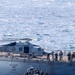 A military maritime operation is underway showing a rigid-hull inflatable boat (RHIB) with armed personnel alongside a larger naval vessel. A military helicopter marked '714' is positioned on the deck of the larger ship, with crew members visible along the ship's railing. A red boarding ladder is deployed on the ship's hull, suggesting a visit, board, search, and seizure (VBSS) or similar naval boarding operation.
