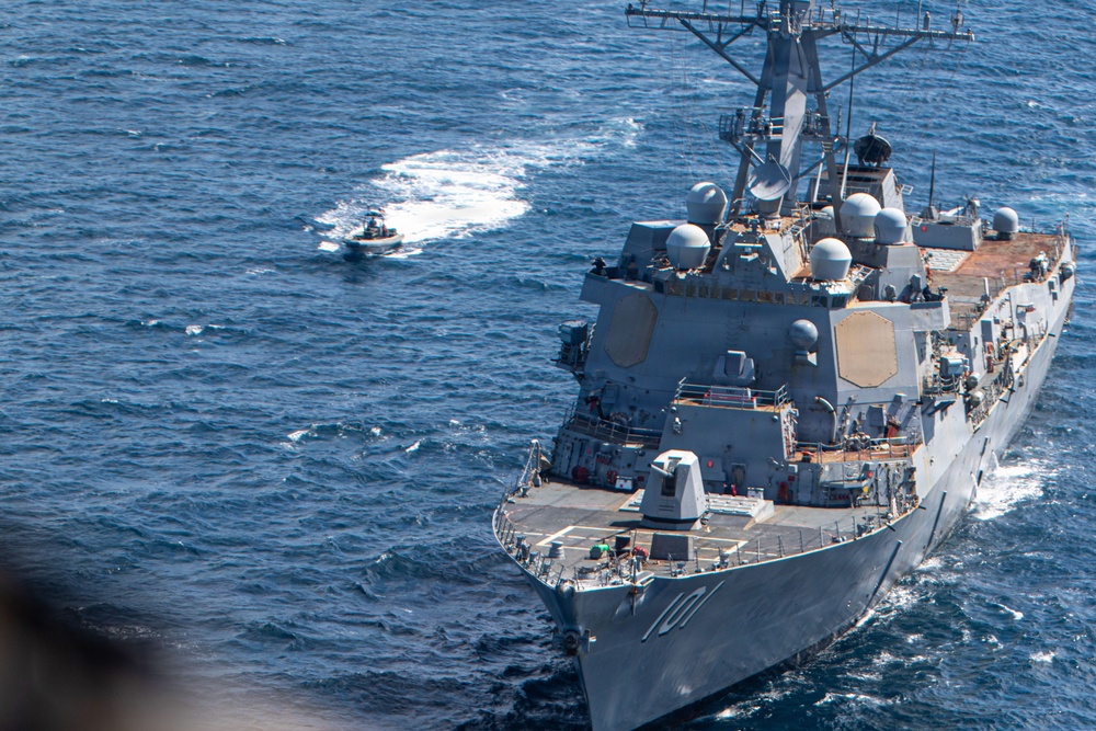 An aerial photograph shows a large US Navy Arleigh Burke-class guided-missile destroyer, hull number 101, underway at sea. A significantly smaller vessel is visible ahead and to the port side of the destroyer, leaving a white wake. The image appears to capture a potential close-approach or intercept scenario between the two vessels.