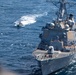 An aerial photograph shows a large US Navy Arleigh Burke-class guided-missile destroyer, hull number 101, underway at sea. A significantly smaller vessel is visible ahead and to the port side of the destroyer, leaving a white wake. The image appears to capture a potential close-approach or intercept scenario between the two vessels.
