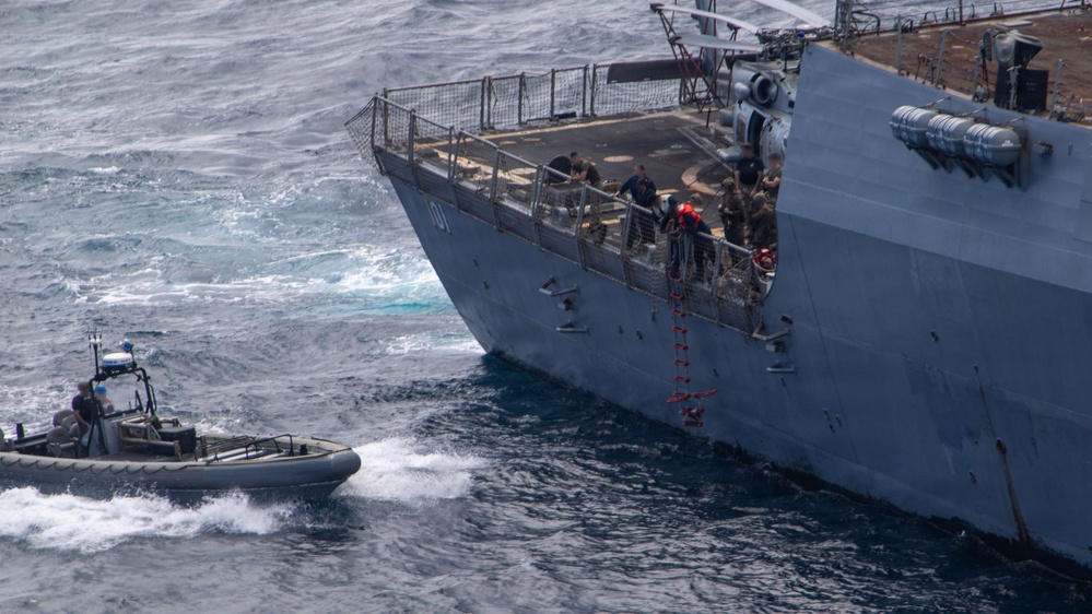 An aerial photograph shows a large gray naval vessel with the hull number '01' partially visible, alongside a smaller rigid inflatable boat (RIB). Personnel are visible on the bow deck of the larger ship, and a rope ladder is deployed over the side. The scene appears to depict a maritime boarding or transfer operation in open, choppy seas.