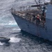 An aerial photograph shows a large gray naval vessel with the hull number '01' partially visible, alongside a smaller rigid inflatable boat (RIB). Personnel are visible on the bow deck of the larger ship, and a rope ladder is deployed over the side. The scene appears to depict a maritime boarding or transfer operation in open, choppy seas.